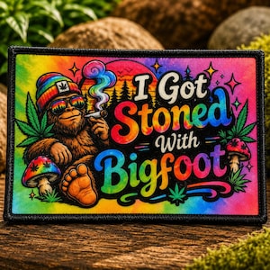 May include: Rectangular patch with a colorful tie-dye background and the text "I Got Stoned With Bigfoot." The patch features a cartoon Bigfoot smoking a cigarette, wearing sunglasses and a beanie with a cannabis leaf.