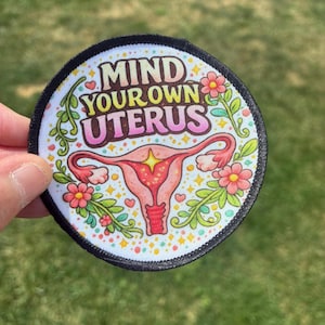 May include: A circular embroidered patch with a black border featuring the text MIND YOUR OWN UTERUS in a gradient font. The design includes a central pink anatomical uterus illustration, surrounded by colorful flowers, green vines, and stars.