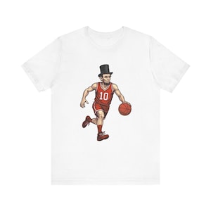 Op de afbeelding: Wit t-shirt met een cartoon illustratie van Abraham Lincoln in een basketbaluniform, dribbelend met een basketbal. De illustratie is in rood, bruin en zwart. Het rugnummer is 10.