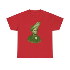 Può includere: T-shirt rosso con un'illustrazione verde di Abraham Lincoln con una pannocchia di mais come cappello. Il design è una rappresentazione umoristica dell'ex presidente americano. La maglietta ha un classico girocollo.