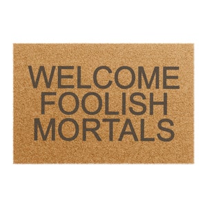 May include: A rectangular doormat made of natural brown coir fiber. The mat features the words "WELCOME FOOLISH MORTALS" in dark gray block letters, creating a welcoming yet humorous entrance.
