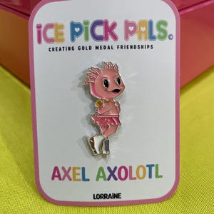 May include: A pink enamel pin featuring a cartoon axolotl on ice skates, with the text "AXEL AXOLOTL" and "ICE PICK PALS." The axolotl wears a pink tutu and has a gold medal. The pin is on a white card with a pink border.