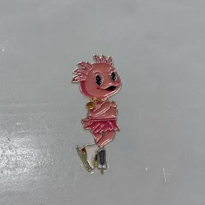 May include: A pink enamel pin depicting a cartoon axolotl figure skater. The axolotl has a pink face, a frilly skirt, and is wearing ice skates. The pin has gold-colored accents.