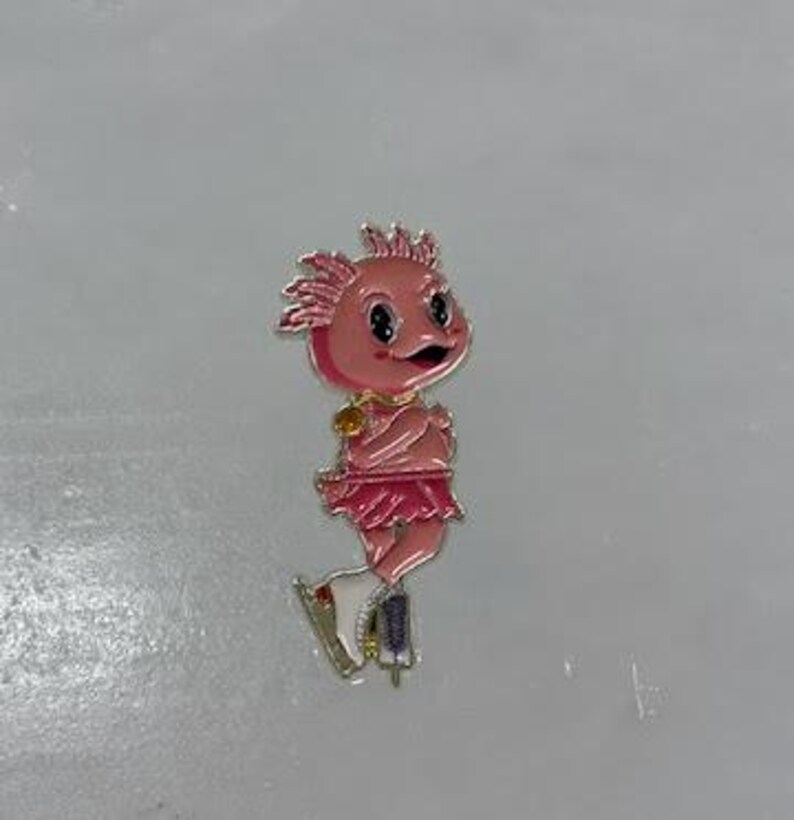 May include: A pink enamel pin depicting a cartoon axolotl figure skater. The axolotl has a pink face, a frilly skirt, and is wearing ice skates. The pin has gold-colored accents.
