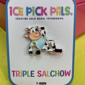 May include: A pin featuring a cartoon cow on ice skates, wearing a blue shirt. The pin is on a white card with the text "ICE PICK PALS" and "TRIPLE SALCHOW". The cow has black and white spots.