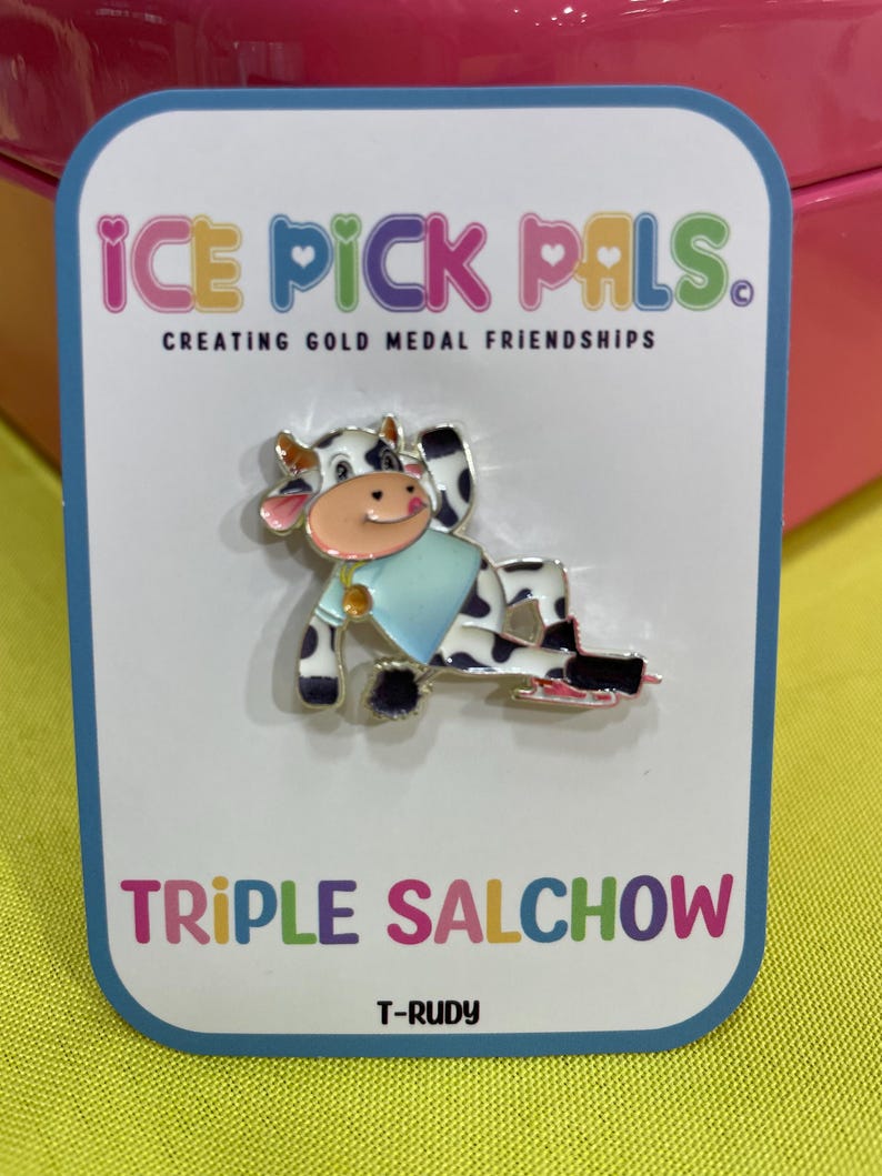 May include: A pin featuring a cartoon cow on ice skates, wearing a blue shirt. The pin is on a white card with the text "ICE PICK PALS" and "TRIPLE SALCHOW". The cow has black and white spots.