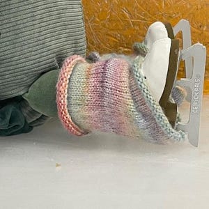 May include: A pair of white ice skates with silver blades, partially covered by a colorful knitted cozy. The cozy features a gradient of pink, purple, and blue hues. The skates are resting on a white surface.