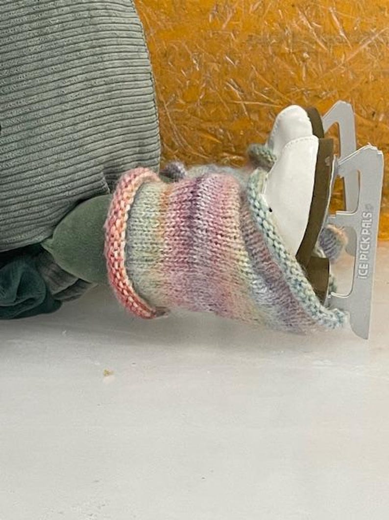 May include: A pair of white ice skates with silver blades, partially covered by a colorful knitted cozy. The cozy features a gradient of pink, purple, and blue hues. The skates are resting on a white surface.