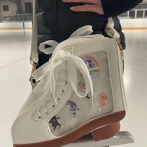 May include: White ice skate-shaped crossbody bag with a clear front panel displaying colorful pins. The bag has a brown sole and gold-colored hardware. A white strap and laces complete the design. The bag is held in front of an ice rink.