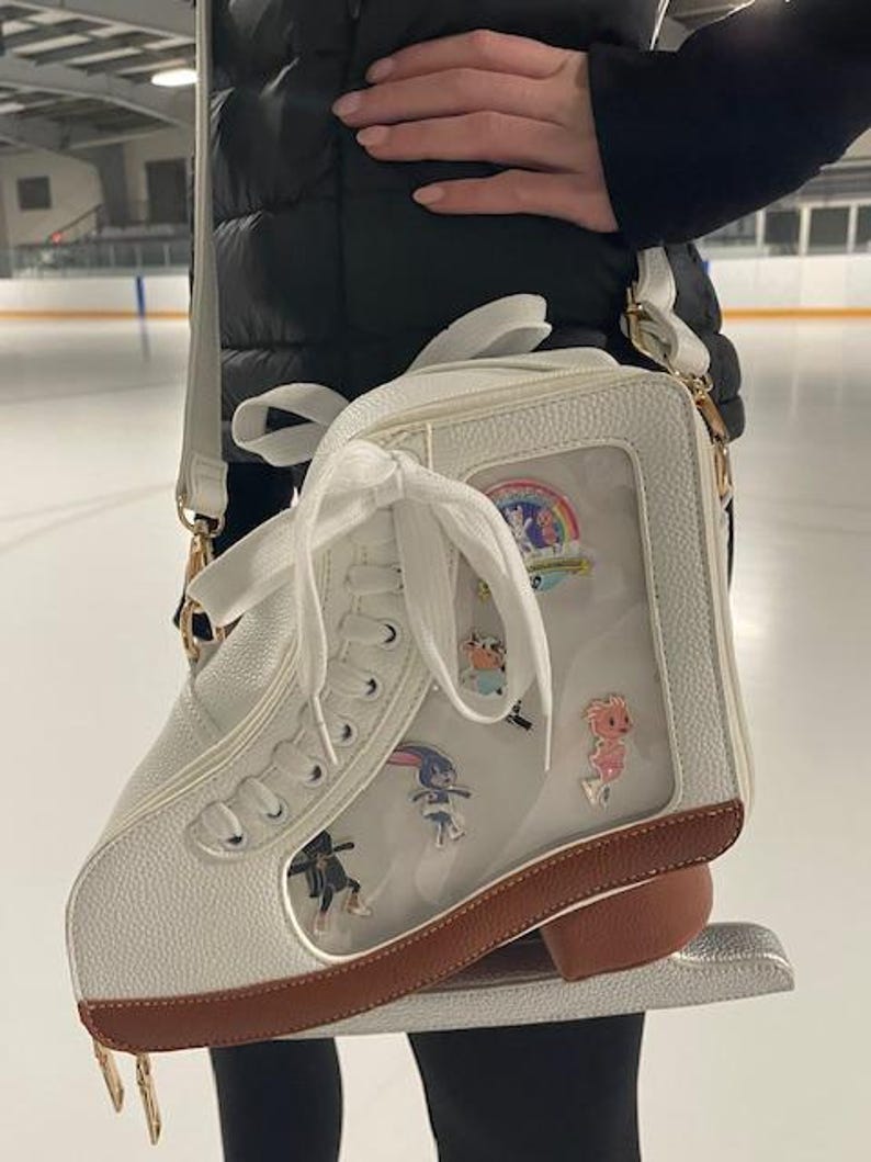 May include: White ice skate-shaped crossbody bag with a clear front panel displaying colorful pins. The bag has a brown sole and gold-colored hardware. A white strap and laces complete the design. The bag is held in front of an ice rink.
