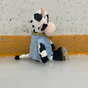 May include: A plush cow toy with black spots, wearing a light blue shirt and black ice skates, sits on an ice rink. The cow has a pink nose and ears, and a yellow collar. The background is a white wall with a yellow border.