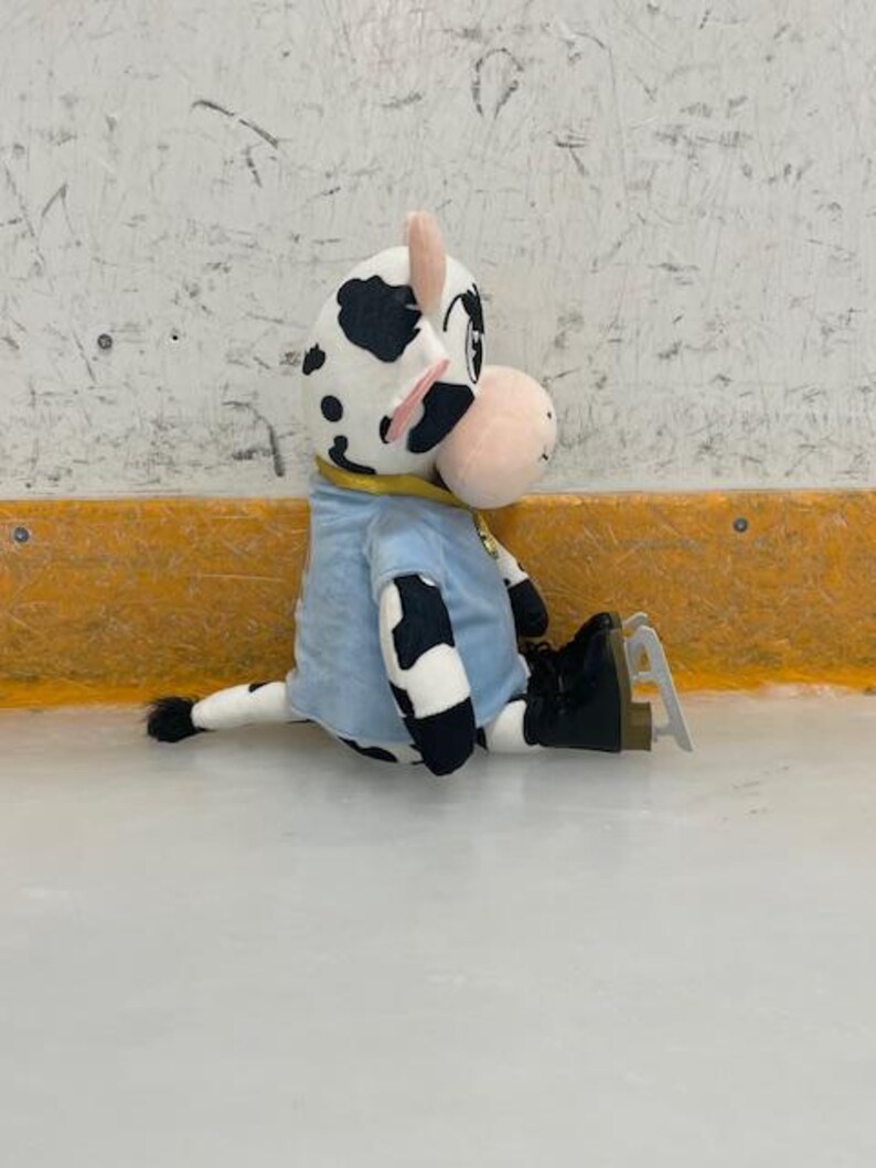 May include: A plush cow toy with black spots, wearing a light blue shirt and black ice skates, sits on an ice rink. The cow has a pink nose and ears, and a yellow collar. The background is a white wall with a yellow border.