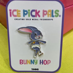 May include: A pin featuring a blue bunny wearing a white skirt and roller skates. The pin is on a white card with the text "ICE PICK PALS" and "BUNNY HOP". The card has a purple border and the name "TAMMY" at the bottom.