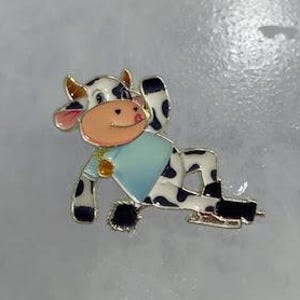 May include: A whimsical enamel pin featuring a cartoon cow in a relaxed pose. The cow has black and white spots, a light blue shirt, and is wearing ice skates. The pin has gold-colored accents and a pink nose and ears.