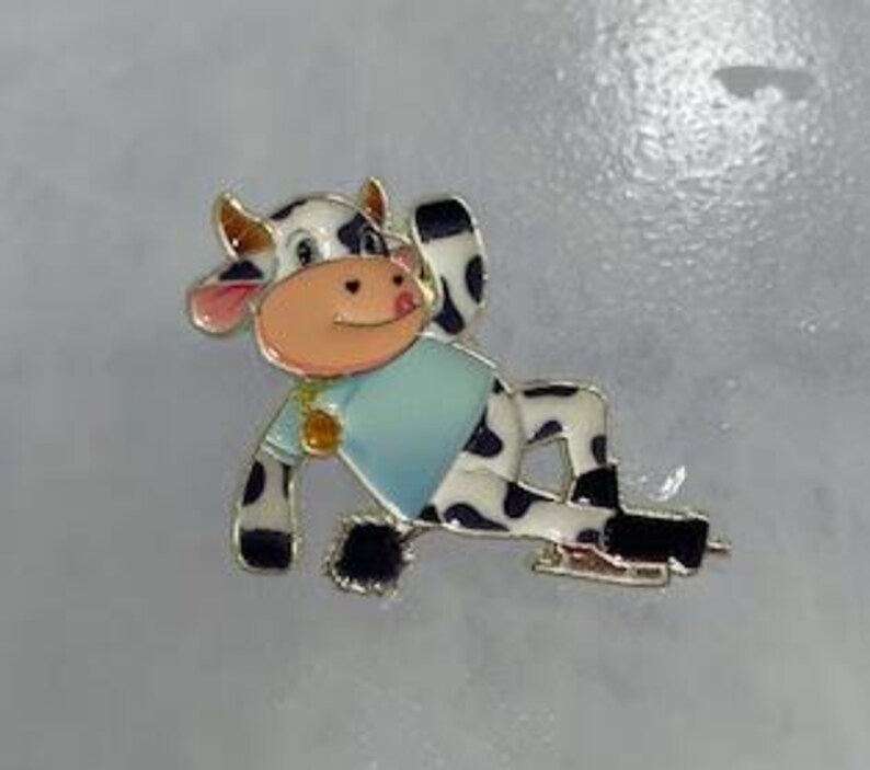 May include: A whimsical enamel pin featuring a cartoon cow in a relaxed pose. The cow has black and white spots, a light blue shirt, and is wearing ice skates. The pin has gold-colored accents and a pink nose and ears.