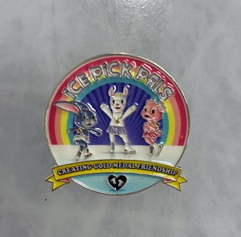 May include: A round enamel pin featuring three cartoon characters against a rainbow backdrop. The pin has the text "ICE PICK PALS" and "CREATING GOLD MEDAL FRIENDSHIP". The characters include a bunny, a llama, and a creature with a pink head.