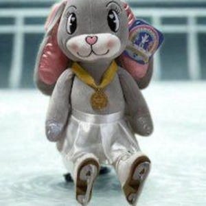 May include: A plush gray bunny wearing a white satin skirt, ice skates, and a gold medal. The bunny has pink inner ears and a pink nose. It is sitting on a blue surface with white lines.