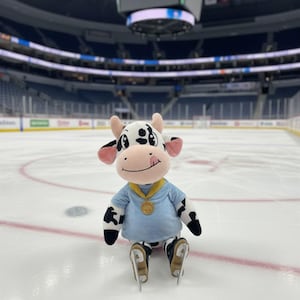 May include: A plush cow mascot wearing a light blue shirt, black and white spotted pants, and ice skates, sitting on an ice rink. The mascot has a gold medal around its neck and is smiling. The background shows an arena with seating.