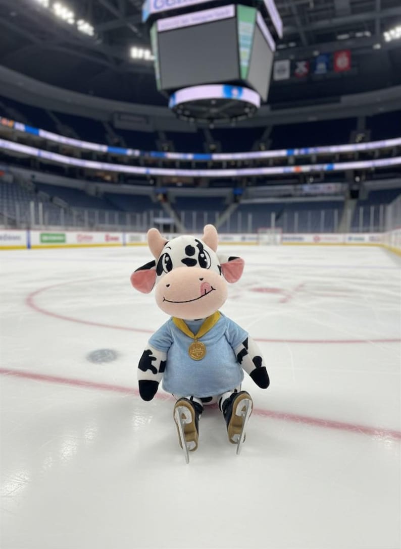 May include: A plush cow mascot wearing a light blue shirt, black and white spotted pants, and ice skates, sitting on an ice rink. The mascot has a gold medal around its neck and is smiling. The background shows an arena with seating.