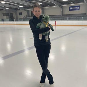 May include: A young figure skater in black athletic wear holds a green plush turtle on an ice rink. The plush toy has a gold medal. The skater is wearing ice skates and is standing on the ice.