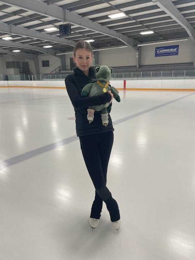 May include: A young figure skater in black athletic wear holds a green plush turtle on an ice rink. The plush toy has a gold medal. The skater is wearing ice skates and is standing on the ice.