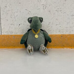 May include: A plush green turtle toy wearing a gold medal and ice skates. The turtle is sitting on an ice rink, with a white and gray background. The toy has a corduroy body and dark green limbs.