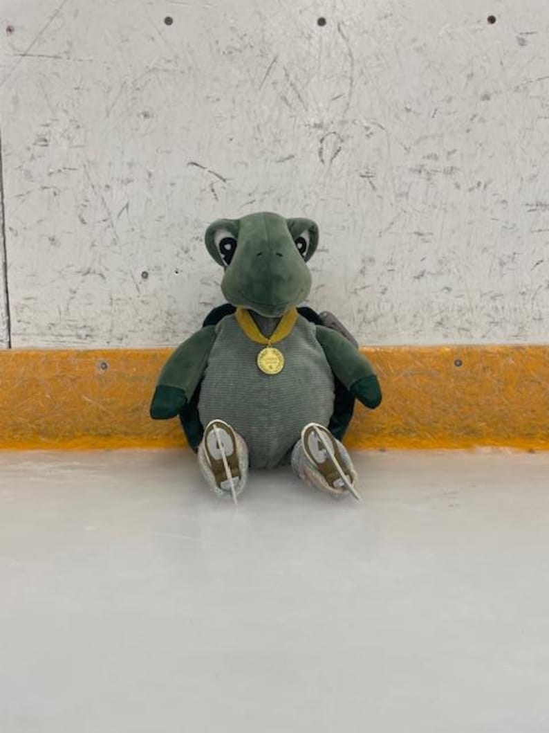 May include: A plush green turtle toy wearing a gold medal and ice skates. The turtle is sitting on an ice rink, with a white and gray background. The toy has a corduroy body and dark green limbs.