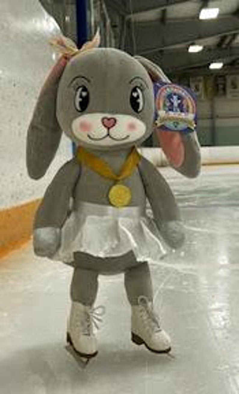 May include: A plush gray bunny wearing a white tutu, ice skates, and a gold medal. The bunny has long floppy ears with pink inner ears and a pink bow. A tag hangs from its ear. The bunny is standing on an ice rink.