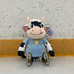 May include: A plush cow toy with black and white spots, wearing a light blue shirt and ice skates. The cow has a pink tongue sticking out and a gold medallion. A tag is attached to the toy.