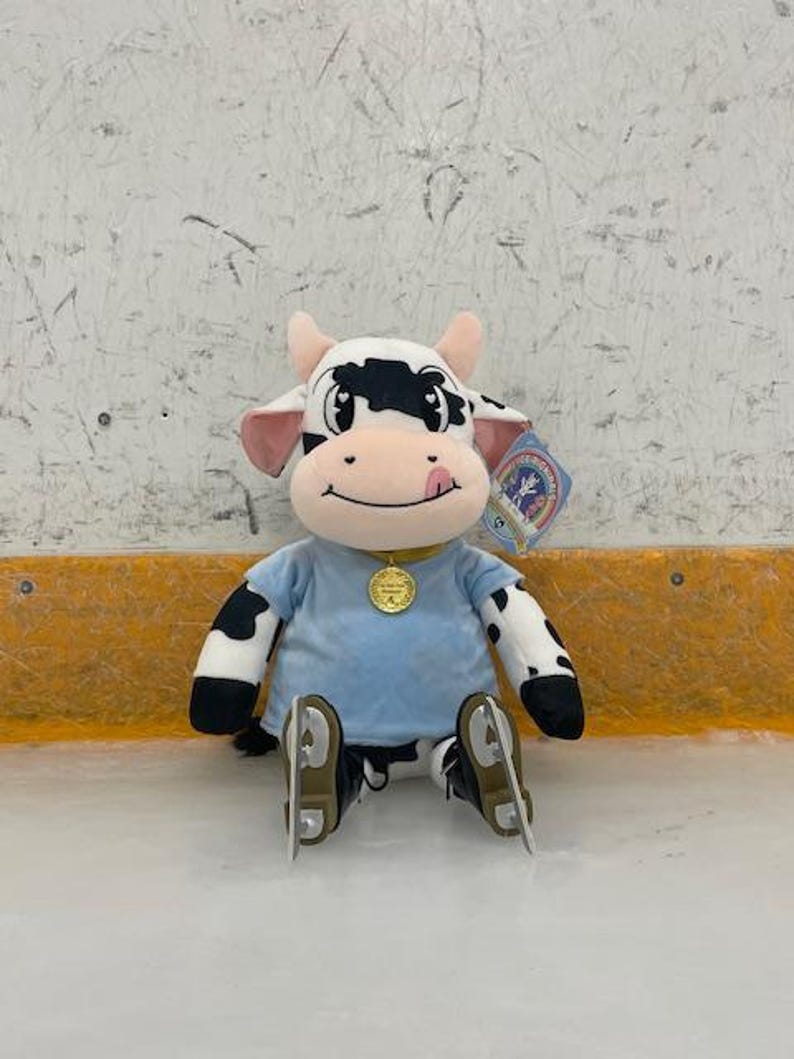 May include: A plush cow toy with black and white spots, wearing a light blue shirt and ice skates. The cow has a pink tongue sticking out and a gold medallion. A tag is attached to the toy.