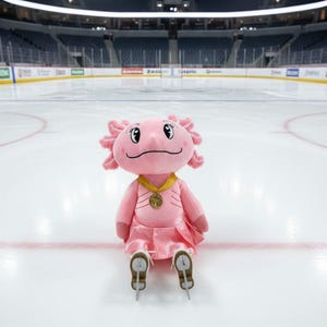 May include: A pink axolotl plush toy wearing a gold medal and a pink dress, sitting on an ice rink. The plush has skates on its feet and is smiling. The background shows an empty hockey arena.