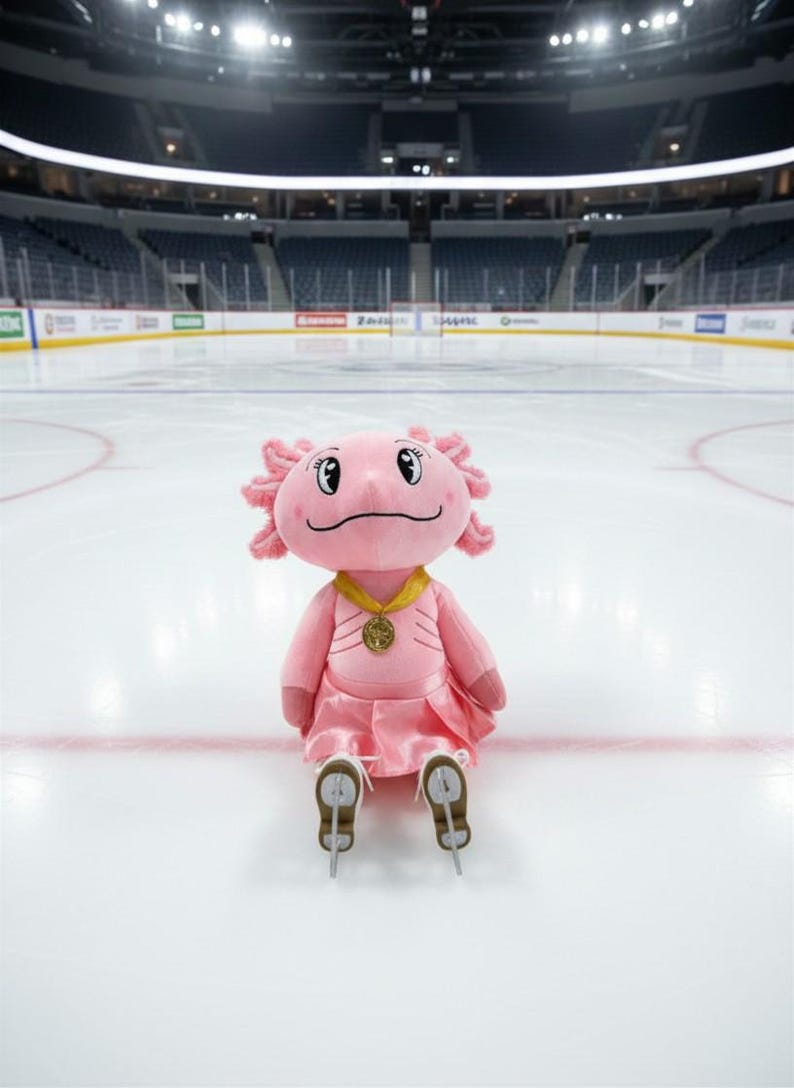 May include: A pink axolotl plush toy wearing a gold medal and a pink dress, sitting on an ice rink. The plush has skates on its feet and is smiling. The background shows an empty hockey arena.