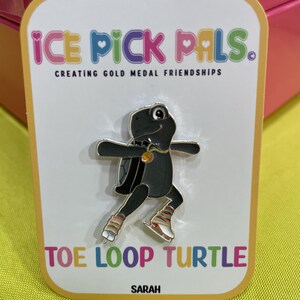 May include: A black turtle pin with silver accents, wearing ice skates and a gold necklace, is featured on a white card. The card reads "ICE PICK PALS" and "TOE LOOP TURTLE". The pin is on a yellow background.