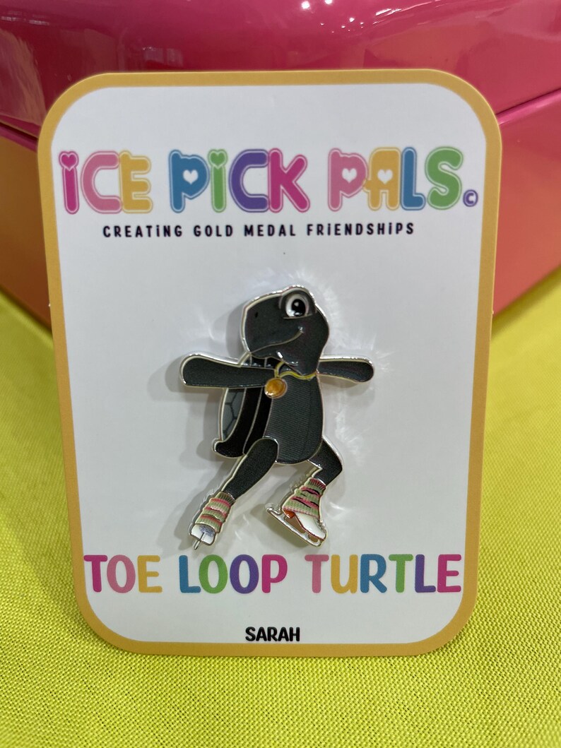 May include: A black turtle pin with silver accents, wearing ice skates and a gold necklace, is featured on a white card. The card reads "ICE PICK PALS" and "TOE LOOP TURTLE". The pin is on a yellow background.