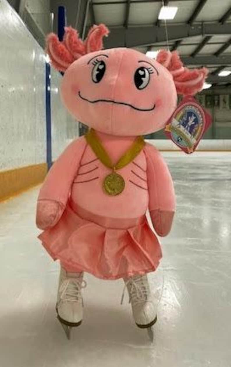 May include: A plush pink axolotl toy wearing a gold medal and a pink skirt, standing on ice skates. The axolotl has a smiling face and decorative gills. A tag is attached to the axolotl.