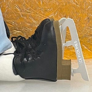 May include: Black ice skate boot with laces and a white blade guard. The blade guard has the text "ICE PICK PALS" printed on it. The boot has a brown sole and is resting on a white surface.