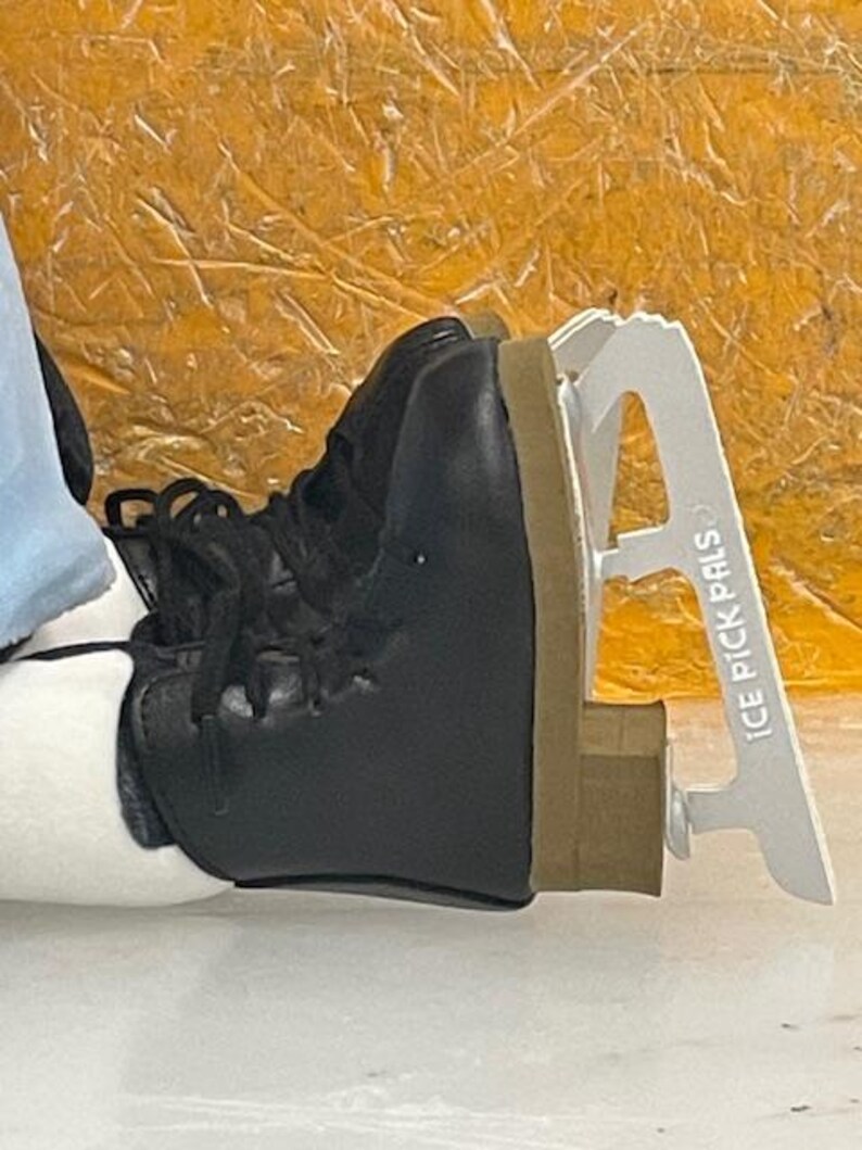 May include: Black ice skate boot with laces and a white blade guard. The blade guard has the text "ICE PICK PALS" printed on it. The boot has a brown sole and is resting on a white surface.