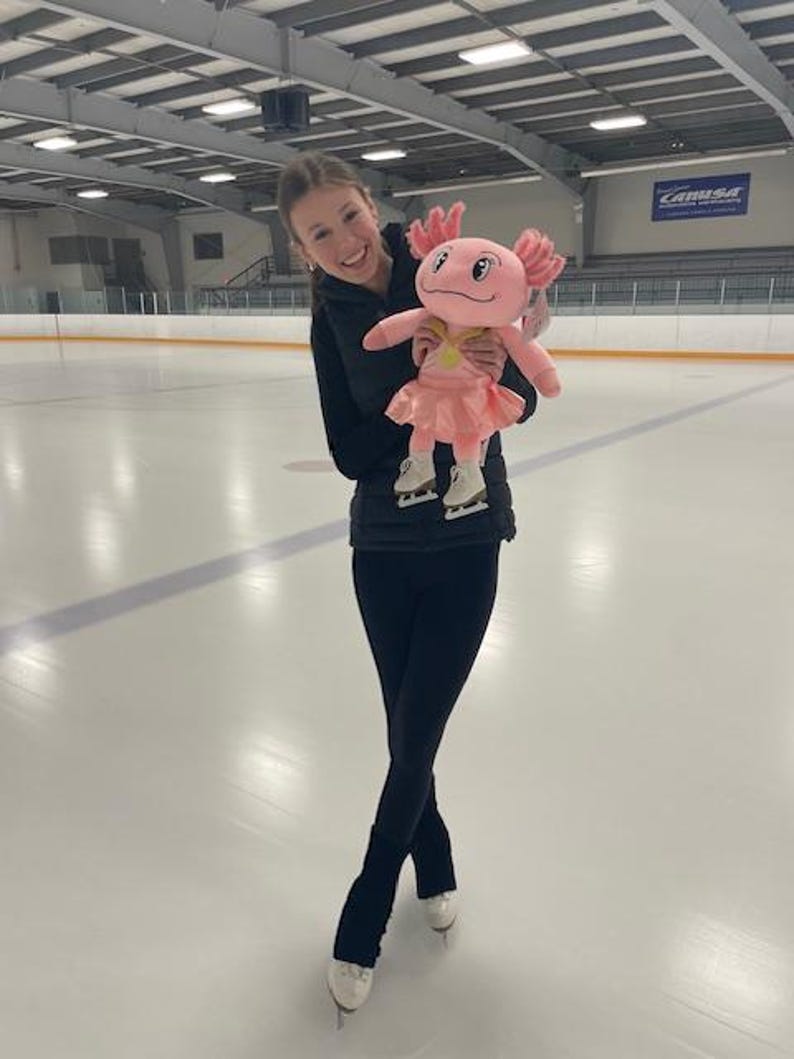 May include: A plush pink axolotl toy wearing a pink skirt and ice skates is held by a person on an ice rink. The axolotl has a yellow necklace and a smiling face. The person is wearing black leggings and ice skates.