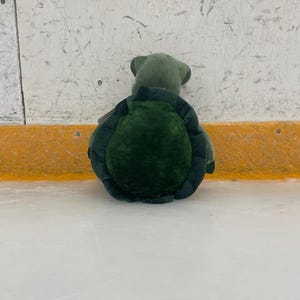 May include: A plush turtle toy, primarily green, with a textured shell and a soft, rounded body. The turtle is positioned on a white surface, with a yellow and white background.