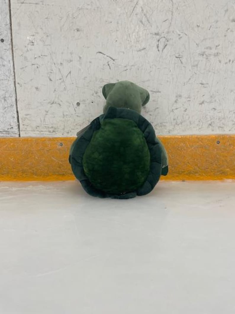 May include: A plush turtle toy, primarily green, with a textured shell and a soft, rounded body. The turtle is positioned on a white surface, with a yellow and white background.