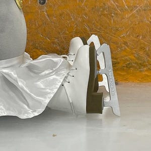 May include: White ice skates with silver blades, labeled "ICE PICK PALS". The skates are laced up and appear to be new. The background is a mix of yellow and white.