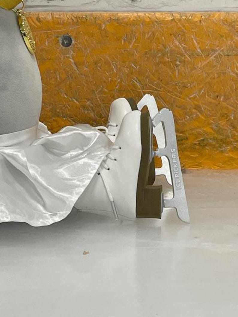 May include: White ice skates with silver blades, labeled "ICE PICK PALS". The skates are laced up and appear to be new. The background is a mix of yellow and white.