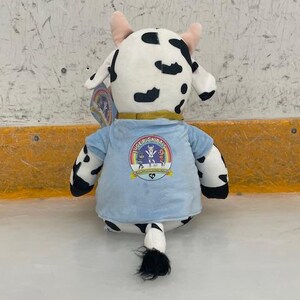 May include: A plush cow toy with black spots, pink horns, and a light blue shirt. The shirt features a colorful logo with the text "ICE PICK PALS". The toy is sitting on a white surface.