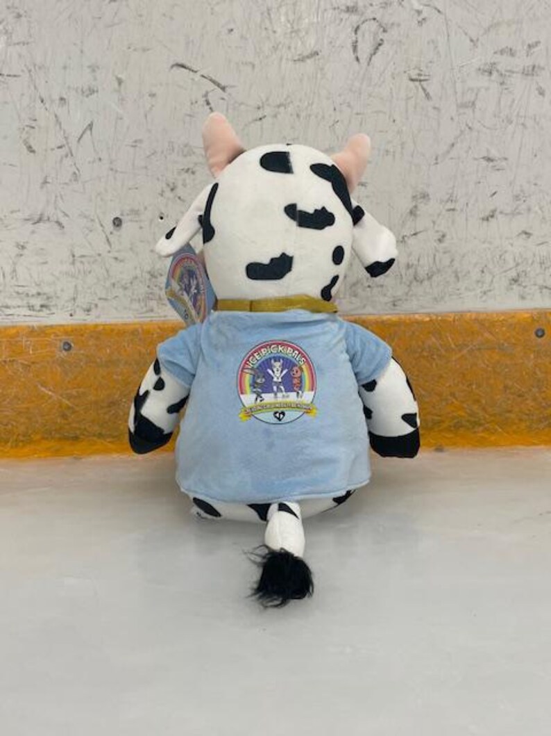 May include: A plush cow toy with black spots, pink horns, and a light blue shirt. The shirt features a colorful logo with the text "ICE PICK PALS". The toy is sitting on a white surface.