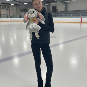 May include: A young person stands on an ice rink, holding a plush bunny toy. The bunny has gray fur, pink ears, and a white tutu. The person is wearing black leggings, a black vest, and ice skates.