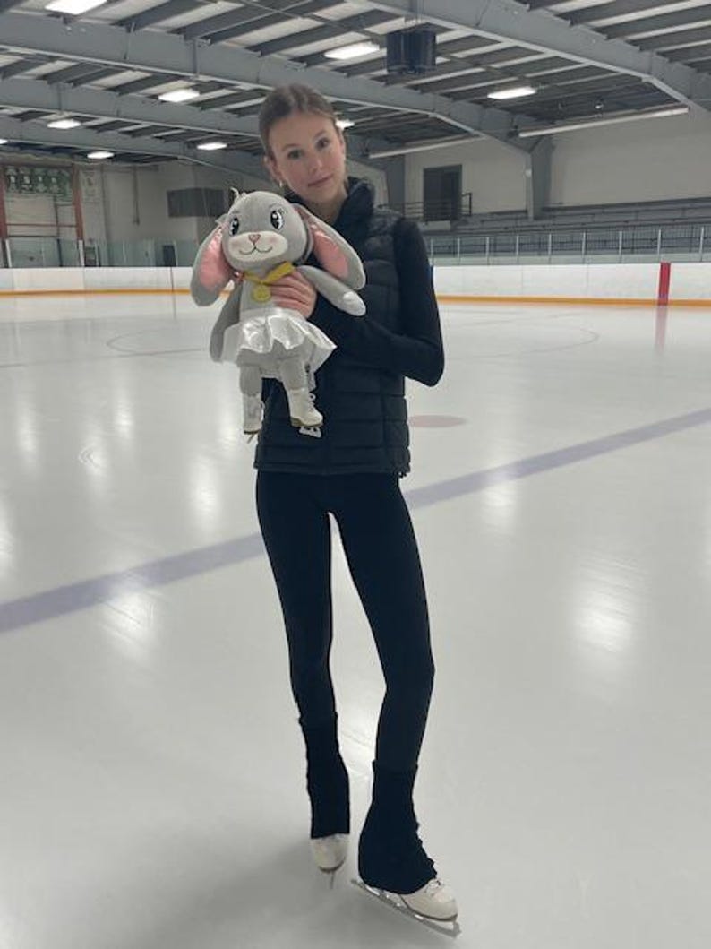 May include: A young person stands on an ice rink, holding a plush bunny toy. The bunny has gray fur, pink ears, and a white tutu. The person is wearing black leggings, a black vest, and ice skates.
