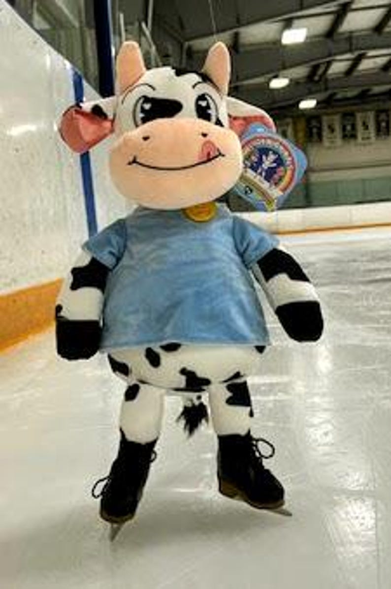 May include: A plush cow toy with black and white spots, wearing a light blue shirt and black ice skates. The cow has pink ears, a smiling face, and a gold tag. It is standing on an ice rink.