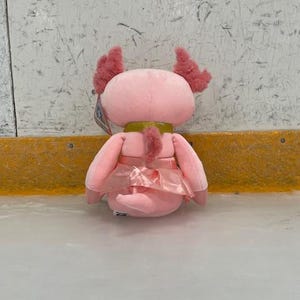 May include: A pink plush axolotl toy with feathery gills and a satin skirt. The stuffed animal has a gold-colored collar and is sitting on a light-colored surface.