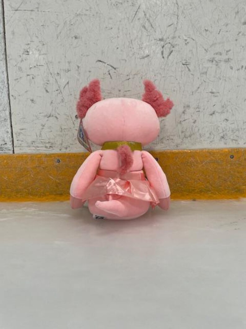 May include: A pink plush axolotl toy with feathery gills and a satin skirt. The stuffed animal has a gold-colored collar and is sitting on a light-colored surface.