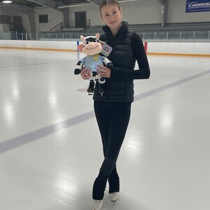 May include: A young person in black athletic wear and ice skates stands on an ice rink, holding a plush cow toy. The cow is white and black with a blue shirt and a yellow collar. The background shows an indoor ice rink.